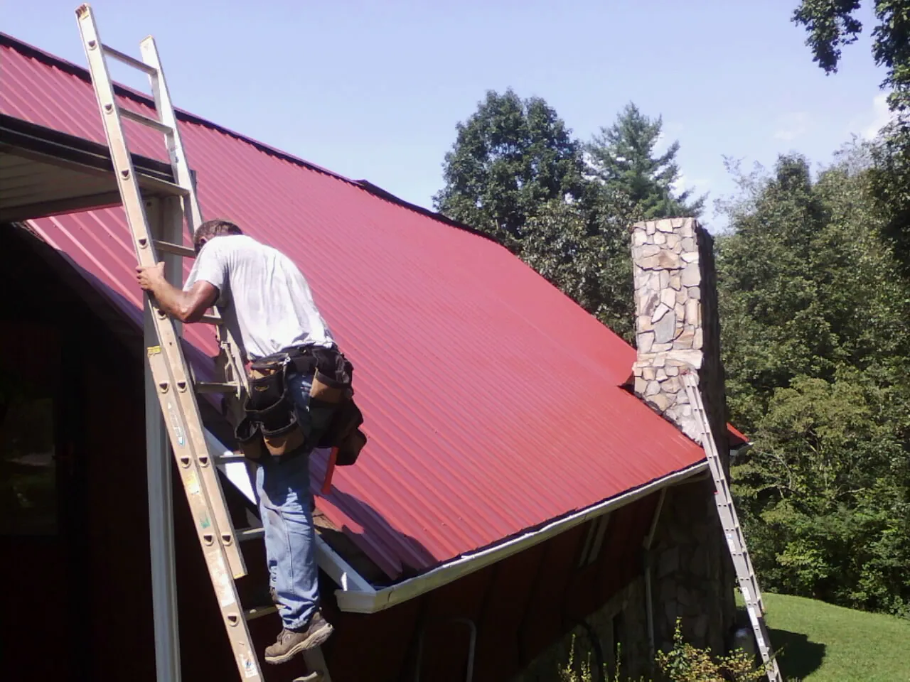Roofer on a ladder beside a red metal roof and stone chimney — Soffit & Fascia Repair by Roofing Brothers in Bourbon